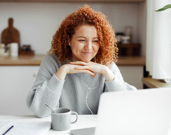 Mujer de mediana edad sonriendo y mirando una pantalla de ordenador con unos auriculares puestos, todo ello en la mesa de su casa